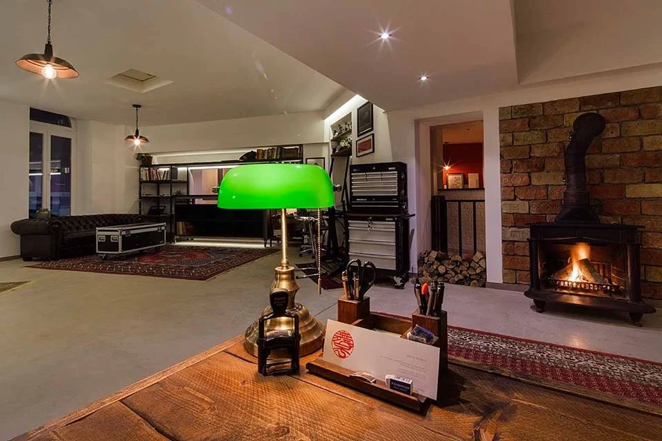 Im Kreativ-Studio der John Ross Group: Schreibtisch mit grüner Bankerlampe, daneben ein Ofen mit Feuer